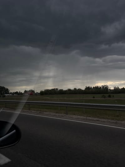 The image depicts a view from a vehicle traveling on a road, likely in a rural area near Balástya, Hungary, although specific landmarks are not visible. The foreground consists of a portion of the asphalt road and a roadside guardrail. Adjacent to the guardrail is a grassy area leading to a line of trees and structures, including what appears to be a red building or vehicle. The sky is overcast with dark, ominous storm clouds dominating the left side of the frame, while shafts of sunlight break through the cloud cover on the right, suggesting a dynamic weather pattern. Part of the car is reflected in the corner. There are no visible people in the frame.