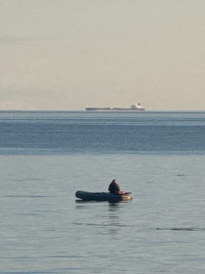 The image depicts a wide expanse of calm water, likely the Gulf of Finland, near Fort Krasnaya Gorka, Russia. The horizon line is clearly defined, separating the muted blue water from a pale, hazy sky. In the middle distance, a large, elongated cargo ship is visible, its silhouette a dark stripe against the lighter sky. Closer to the foreground, a small, dark inflatable boat is floating on the water. A single person, seen from behind, is seated in the boat, appearing to be fishing or simply observing the surroundings. The water is mostly smooth, with subtle ripple patterns that suggest a gentle breeze. The overall atmosphere is one of quiet solitude and natural beauty.