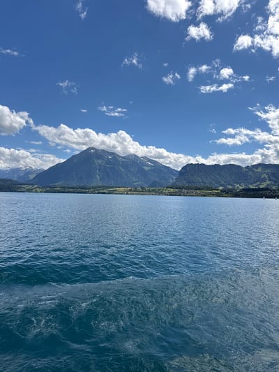 The image displays a wide expanse of blue water with ripples, possibly a lake, in the foreground. In the background, there is a mountainous landscape covered in greenery and some snow-capped peaks. A small town or village is nestled at the base of the mountains, with scattered houses and buildings. The sky is a vibrant blue, dotted with white, fluffy clouds. The scene suggests a scenic natural environment, likely in a mountainous region of Switzerland, given the mention of Hünibach. There are no people or specific actions visible in the image. The overall impression is one of tranquility and natural beauty.