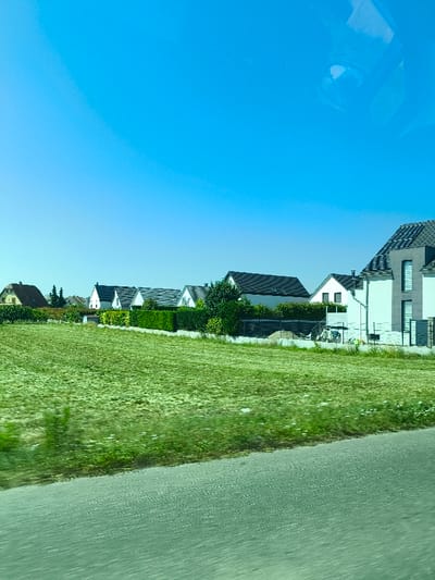 A bright blue sky overlooks a row of houses along a road. A field of green grass stretches between the road and the houses, and the houses are various shapes and sizes with dark roofs and light-colored walls. Some have small yards or fences, and trees dot the landscape. The composition has an impressionistic, slightly dreamy feel.