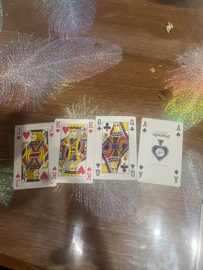 Four playing cards are laid out on a wooden table with a decorative, glittery overlay. The cards, from left to right, are the Jack of Hearts, the King of Hearts, the Queen of Clubs, and the Ace of Spades. The Ace of Spades card has text in black that reads "PIATNIK VIENNA" and "MADE IN CHINA". There are a few small crumbs and a brown nut on the table. The scene is not located in the city of Rosenovo, Bulgaria.