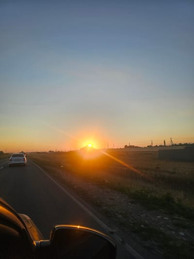 The image shows a sunset over a rural landscape in the village of Kokshayak, Kazakhstan. The sun is low on the horizon, casting a warm, orange glow across the scene. A road runs diagonally across the image, with two cars driving on it. The car in the foreground is a white sedan, and the car behind it is a dark-colored SUV. The road is flanked by dry grass and sparse vegetation. In the distance, there are a few houses and power lines. The sky is a clear blue, with a gradient of orange and yellow near the horizon. The image appears to have been taken from inside a moving vehicle, as evidenced by the reflection in the side mirror and the slight motion blur. The overall mood of the image is peaceful and serene.