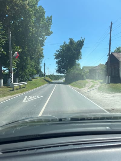 The image shows a view from inside a vehicle driving on a paved road in Yadrin, Russia. The road is flanked by trees and a grassy verge on both sides. On the left side, there is a utility pole with a small Russian flag attached, and a white metal guardrail. Further down the road on the left, a white fence with trees behind it is visible. On the right side of the road, there is a dirt path leading to several wooden buildings, one of which appears to be a house with ornate window frames. The sky is clear and blue. The road itself has white lane markings, and a triangular road sign depicting a child crossing is painted on the pavement in the foreground. The dashboard of the vehicle is visible at the bottom of the frame. The overall impression is a rural or semi-rural setting on a sunny day.