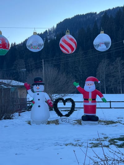 The image displays an outdoor winter scene in Gstaad, Switzerland, featuring Christmas decorations. In the foreground, a snow-covered ground extends across the frame. Two large inflatable figures stand centrally: an inflatable snowman on the left, wearing a black top hat with a red band, a green and red striped scarf, and red mittens, with its right arm raised in a wave. To its right stands an inflatable Santa Claus, dressed in a red suit with white trim, a black belt, green gloves, and a red hat, with its left arm raised in a wave. Between the two figures, a dark green heart-shaped wreath is positioned on a stand, bearing a white sign that reads "Weihnachts Paradies" (Christmas Paradise).

Above these ground-level decorations, a horizontal wire stretches across the upper portion of the frame, from which several large, spherical Christmas ornaments are suspended. From left to right, a partially visible green ornament with red and white decorations, a transparent ornament containing a festive scene with a reindeer and gifts, a red and white striped ornament, and another transparent ornament containing a snowman figure are visible.

The mid-ground features a sparse arrangement of bare deciduous trees and shrubs. A wooden fence runs horizontally behind the inflatable figures, separating the decorated area from a wider snow-covered field. In the background, a dense coniferous forest covers the lower slopes of a mountain, which itself shows patches of snow. The sky above is clear and light blue, indicating daylight.
