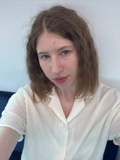 The image shows a medium shot of a young woman with light skin and medium brown, wavy, shoulder-length hair. She has blue eyes and is looking directly at the camera with a slight smile. She is wearing a cream-colored, short-sleeved shirt with a collar and buttons. The background is a plain white wall, and a blue surface is visible on the left side of the image. There is no discernible location in the city of Yangju, South Korea.