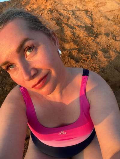 A woman with blonde hair and blue eyes is photographed from a high angle, looking up towards the camera. She is wearing a pink and navy blue swimsuit with the logo "JOSS" visible on the chest. Her left arm is resting on her chest, and her right arm is out of frame. The background consists of sandy terrain, illuminated by warm, golden sunlight, suggesting it is either sunrise or sunset. The environment appears to be outdoors, possibly a beach or a sandy embankment. The location is identified as Sapunov Bor, Russia.