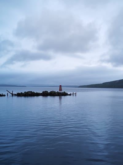 The image presents a serene and expansive landscape dominated by a large body of water under a heavily overcast sky. In the mid-ground, a small, rocky island or breakwater stretches across the water, constructed from dark rocks and weathered wooden debris. A prominent feature on this island is a striking red and white striped structure, resembling a navigational beacon or a small lighthouse, which stands vertically with distinct red and white bands.

Distant forested shores or hills frame the far edges of the water on both the left and right, receding into a hazy horizon and suggesting a vast lake or wide river. The sky is completely covered by thick, grey clouds, indicating an overcast day and casting a soft, diffused light over the entire scene. The water surface is a deep blue, textured by gentle ripples that reflect the subdued light of the sky.

No people or explicit activities are visible, contributing to a tranquil and solitary atmosphere. The overall impression is one of calm and natural beauty on a cloudy day. Given the context, this scene likely depicts Lake Onega near Medvezhyegorsk, Russia. There is no visible text in the image.