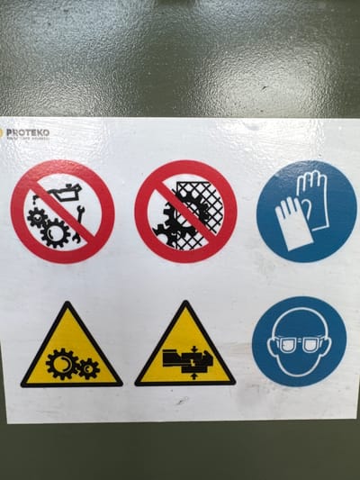 The image shows a sign with multiple safety pictograms, likely affixed to a piece of machinery. The sign is white with black and red graphics.
At the top left, there is a "PROTTEKO" logo.
In the top row, there are three circular prohibition signs with red borders and red diagonal lines. The first shows an oil can and gears, indicating no lubrication or maintenance involving these parts. The second shows gears behind a grid or cage, suggesting that moving parts are protected and should not be accessed. The third, on the right, is a blue circle with a white outline of gloves, indicating the mandatory use of hand protection.
In the bottom row, there are two yellow triangular warning signs with black borders and graphics. The first shows two meshing gears, possibly warning of moving parts or entanglement hazards. The second shows a representation of crushing or pinching hazard with arrows indicating movement towards the center. To the right of these, there is another blue circular sign with a white outline of a face wearing safety glasses, indicating mandatory eye protection.
The sign is placed on a dull, textured surface which appears to be green, possibly a wall or the exterior of equipment. The lighting suggests an indoor environment with overhead light.
This image was taken in Mejaniga, Italy.