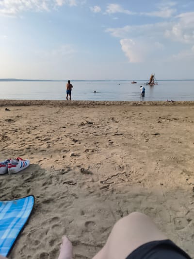 This is a photograph taken at the beach in Gubernskoe, Russia. The foreground features a sandy beach with a blue and white checkered mat and a pair of white athletic shoes with red stripes. In the background, several people are visible in the shallow water. One man stands waist-deep, facing away from the camera. Further out in the water, a floating structure with a slide is present, and other individuals are scattered around the area. The sky above is a pale blue with scattered clouds.