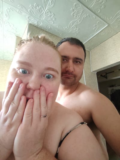 The image shows a selfie taken indoors, likely in a residential setting, with two individuals visible. A woman in the foreground, with fair skin and freckles, is looking at the camera with a surprised expression, her hands covering her mouth. She is wearing a black and white patterned top, and a silver ring with a dark stone is on her left ring finger. Behind her, a man with dark hair and stubble is also looking towards the camera with a neutral expression. The background shows a textured ceiling with floral patterns and a wall with a light beige textured finish. The overall lighting suggests an indoor environment, possibly a bathroom or bedroom. There is no information in the image to suggest the location is Красная Поляна, Russia.