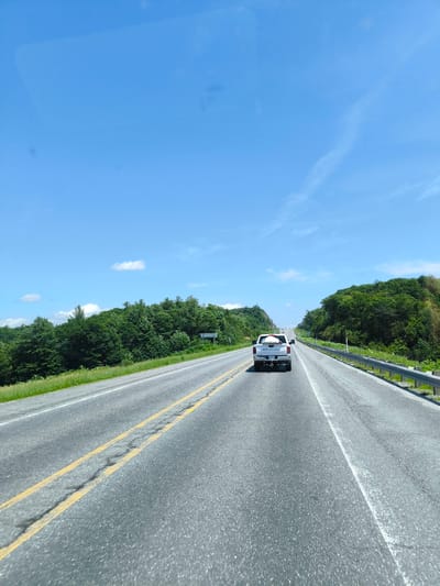 The media file depicts a bright, sunny day on a highway with a clear blue sky and scattered white clouds. The road is paved and has yellow double lines marking the lanes. Lush green trees line both sides of the highway, with one side appearing to be a steeper incline covered in dense foliage. A silver pickup truck with a white object in its bed is traveling on the highway. A metal guardrail is visible along the right side of the road. The scene suggests a scenic drive through a rural or semi-rural area. The time of day is midday, indicated by the bright sunlight and clear sky. The location is identified as Papineauville, Canada. No specific individuals are clearly discernible, and no text is visible in the image.