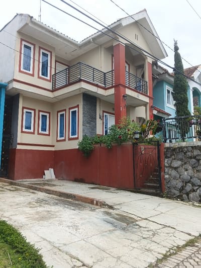 The scene depicts the exterior of a two-story residential structure in Pô Kô, Vietnam, observed under an overcast sky. The primary building, positioned centrally, features a beige upper façade with a gabled roof and a dark red lower half, including supporting columns and a perimeter wall. White-framed rectangular windows with visible blue interior coverings are present on both floors, totaling at least seven. The second floor includes a prominent front balcony and a partial side balcony, both equipped with black horizontal metal railings. A covered entrance porch is located on the ground floor, featuring a white door and a large dark red support column. The number "2B" is affixed to one of the columns near the entrance and also below a second-story window.

To the immediate left, a portion of another blue-painted structure and a dark metal gate are visible. To the right, an adjacent two-story house, painted teal with a brown tiled roof, is partially visible, fronted by a tall, slender evergreen tree and a black metal fence.

In the foreground, a concrete driveway or pathway slopes upward towards the houses, displaying visible cracks and minor discoloration. A low concrete curb runs across the pathway. Small patches of green grass line the very edges of the foreground. Along the base of the central house's dark red wall, several rectangular light-colored construction materials are stacked. A black ornate metal gate provides pedestrian access to the central property, leading to unseen steps. A stone retaining wall constructed from irregular grey rocks is visible to the right of the central property's gate. Multiple black utility lines are strung across the upper portion of the frame. No individuals or active interactions are present in the image.