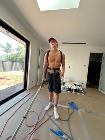 A shirtless man in a cap cleans a room, operating a floor cleaner. He wears shorts, white socks, and a harness. The room has white walls, modern black-framed windows, and a tan floor. A doorway leads to another room with construction materials. Cleaning supplies, including a brush and dustpan, are on the floor, along with an extension cord. Sunlight streams in through a window.