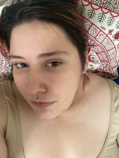 A person is lying down, possibly in bed, with their head on a pillow. The pillow has a red and white patterned fabric with what appear to be stylized floral or leaf motifs. The person's face is turned towards the camera, with their eyes open and looking directly at the viewer. Their hair is dark brown with some reddish highlights. They are wearing a light-colored tank top. The image appears to be a close-up, self-taken photograph. The specific location in Коробицыно, Russia cannot be determined from the image.