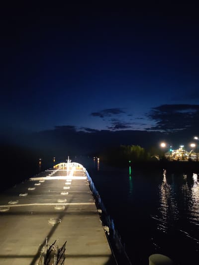The image depicts a nighttime view from the deck of a large ship or barge. The deck is illuminated with a series of lights. The ship is traveling along a waterway. On the right side of the waterway, there is an industrial area with brightly lit buildings, storage structures, and a green light. The sky is a deep blue, with some clouds near the horizon. It could be in or near Александровское, Russia.