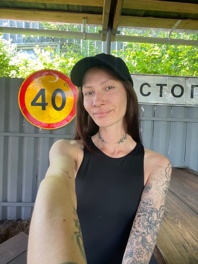 A woman stands in front of a railroad stop in Doma 45 Kilometr, Russia. She is wearing a black tank top and a black hat, and has tattoos on her left arm and neck. She is holding up a circular yellow and red speed limit sign with the number 40 on it. Behind her, a sign reads "CTO." The environment consists of trees and a building with metal and wooden structures.
