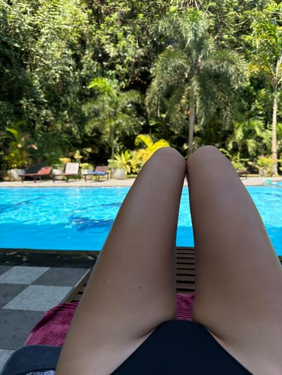 A person is sitting by a pool, with their legs in the foreground. They are wearing a dark swimsuit. The bright blue pool water is visible behind the legs, with palm trees and other greenery in the background. The pool area features lounge chairs and the ground is tiled.