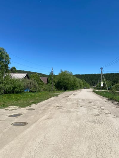 The image depicts a dirt road in the village of Bolshoy Lug, Russia, on a clear, sunny day. The road is unpaved and shows signs of disrepair, with numerous potholes and patches of cracked asphalt. To the left of the road, a grassy embankment is visible, dotted with yellow wildflowers and a few puddles. Beyond the embankment, several houses are partially obscured by lush green trees and bushes. The houses appear to be of traditional wooden construction, with one having a white corrugated metal roof and another a darker, possibly wooden, roof. Power lines run overhead, supported by a wooden pole on the right side of the road. A white car is seen driving away in the distance, kicking up a faint dust trail. The road curves gently to the right, leading towards a dense forest that forms the backdrop of the scene. The sky is a vibrant, cloudless blue.