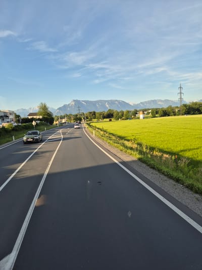 The image captures a view of a road in Muntigl, Austria, under a clear blue sky with scattered clouds. The road, paved with asphalt, has white lane markings. On the left side of the road, there is a grassy area with trees and buildings, including a sign that reads "LINGHAMO" and another building with "LINGHAMO" signage. A green car is driving on the road, and further ahead, two other cars are visible.

To the right of the road, a vibrant green field stretches towards a line of trees. In the background, a range of mountains is visible, with a tall electrical pylon standing prominently in the mid-ground. The sun casts shadows across the road and the field, suggesting it is either late afternoon or early morning. The overall scene depicts a rural or semi-rural environment on a clear day.