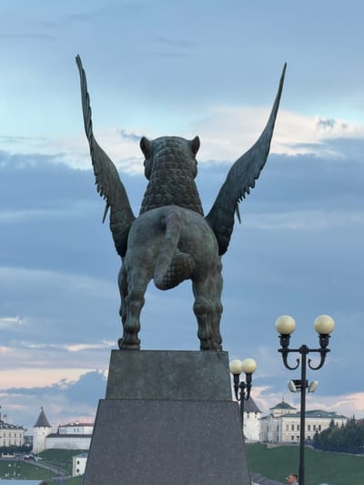 This media file captures a bronze sculpture of a winged creature, possibly a griffin or a dragon, positioned outdoors against a sky with scattered clouds. The sculpture is viewed from behind, showcasing its detailed wings, feathered texture, and powerful hindquarters. It stands on a large, dark grey stone pedestal. In the background, a street lamp with two white globes is visible. Further in the distance, architectural structures that appear to be historical buildings with white facades, green roofs, and turrets can be seen. The lighting suggests it is either early morning or late afternoon, with a soft, diffused light illuminating the scene. The overall atmosphere is calm and serene. There is no visible text in the image. The location context is provided as Yanga-Yul, Russia.