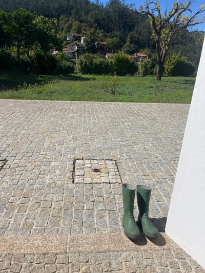 The image displays an outdoor scene in Oliveira de Frades, Portugal, under clear blue skies. The foreground is dominated by a light-toned cobblestone pavement, characteristic of calçada portuguesa. Two dark green rubber boots are positioned upright on this pavement in the lower right, casting distinct shadows to their left. A white vertical surface, likely a wall or column, occupies the extreme right edge of the frame. Embedded in the cobblestones near the center are two rectangular metal utility covers, one with a circular opening. Beyond the paved area, a strip of mown green grass extends across the midground. The background consists of a verdant hillside, densely covered with trees and shrubs. Several white-walled houses with red-tiled roofs are visible scattered among the foliage on the hillside. A large, bushy tree stands on the left side of the midground, while a more slender tree with sparse leaves is on the right. Bright daylight illuminates the scene, generating crisp shadows.