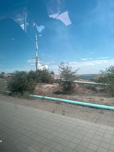 The image depicts a wide shot of a dry, somewhat barren landscape under a bright blue sky with scattered clouds. In the mid-ground, a tall, white, abstract sculpture resembling a rocket or a stylized human figure stands prominently. The sculpture has outstretched arms and a pointed top. Surrounding the sculpture are sparse trees and bushes, with dry, brown earth and some rocky terrain.
In the foreground, a large, teal-colored pipe runs horizontally across the lower part of the frame. The ground in the immediate foreground appears to be a paved surface with a textured pattern, possibly a road or walkway. Further in the background, a body of water, likely a lake or reservoir, is visible, with more distant hills and power lines stretching across the horizon. Reflections of the sky and possibly the interior of a vehicle are visible on a shiny surface in the upper portion of the image, suggesting the photo was taken from inside a car or bus.
The location is identified as Jezqazğan, Kazakhstan. This specific sculpture is likely associated with the history of space exploration in the region, as Jezqazğan is near the Baikonur Cosmodrome. The arid environment and the presence of industrial infrastructure like pipes and power lines are characteristic of this part of Kazakhstan. No people are visible in the image.