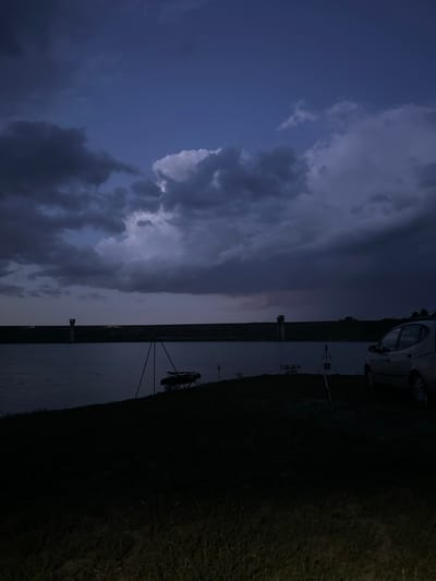 The image depicts a landscape at dusk in Trastikovo, Bulgaria. Dominating the left side is a cloudy sky with varying shades of blue and grey, suggesting an approaching storm. A body of water bisects the image horizontally. The right side includes a dark, grassy area, and possibly a car parked nearby. Further along the water's edge are structures that appear to be related to electrical infrastructure, including towers and poles.