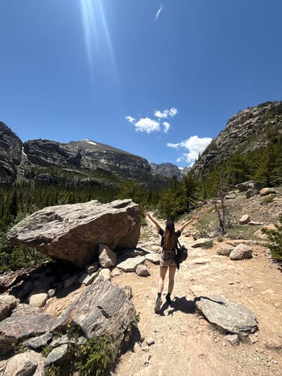 The image depicts a young woman from behind, standing on a dirt trail in a mountainous region, with her arms outstretched towards a clear blue sky. She is wearing a dark top, leopard-print shorts, and dark shoes, with a backpack slung over her left shoulder. The trail is rocky and uneven, with large boulders scattered throughout the landscape. In the background, a dense forest of evergreen trees covers the slopes of steep, rocky mountains, with patches of snow visible on the higher peaks. The sun is shining brightly, casting long shadows on the ground and creating sunbeams in the sky. The location is likely in or around Allenspark, Colorado, given the characteristic Rocky Mountain scenery, which is known for its rugged terrain and pine forests.