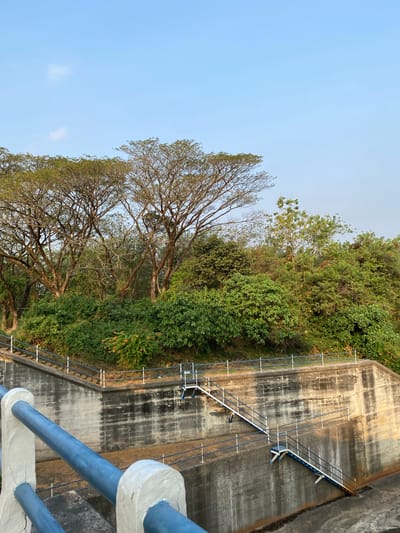 The image shows a concrete dam structure with stairs leading up its side. Lush green trees and foliage cover the top of the dam. The sky is a clear blue with a few wispy clouds. In the foreground, a blue railing with white supports is visible. The scene appears to be an outdoor location, possibly a park or recreational area. The location is identified as a dam in Bandungan, Indonesia.