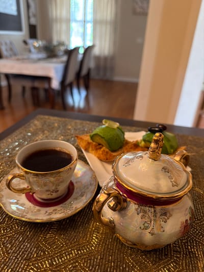 A close-up shot captures a coffee cup and a sugar bowl on a decorative beaded placemat. The coffee cup, filled with dark coffee, sits on a saucer with a red rim and gold trim. The sugar bowl, also adorned with gold accents and intricate patterns, is placed to the right of the cup. In the background, out of focus, is a dining table with chairs, suggesting a residential setting. The overall scene evokes a morning breakfast or a quiet moment with coffee. The location is identified as Riverview, United States of America.