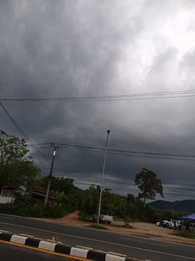 The photograph depicts a street scene in Pak Phraek, Thailand, under a sky heavy with dark, ominous clouds. In the foreground, a two-lane road with a black and white curb borders the asphalt. On the left, a white house is partially visible behind dense foliage. The middle ground contains a row of lush green trees, interspersed with utility poles carrying a web of electrical wires. In the background, a distant range of hills or mountains is discernible through the overcast atmosphere. Several vehicles, including pickups and vans, are parked in a dirt area adjacent to the road on the right side. A lamppost stands prominently amidst the trees near the center of the image. There is no discernible human presence in the frame.