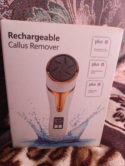 The image displays the front of a product box for a "Rechargeable Callus Remover." The box is primarily white with text and graphics. The product itself, a white and rose gold electric callus remover with a digital display, is partially visible on the right side of the box. The box features several "plus" icons with accompanying text highlighting features such as "Three different roller to customize your results," "Fast and Slow Gear Design," and "Wet&dry use, easy for clean and more comfortable." Water splash graphics are also present on the lower portion of the box. The box is resting on a textured surface, possibly a blanket, with a leopard print pattern. The background shows a patterned textile, likely a blanket or upholstery. The image does not provide any information that would allow for localization in Monastirische, Russia.