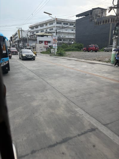 The image, taken in Dagupan, Philippines, depicts a street scene with several vehicles and buildings. The perspective is from inside a vehicle, looking forward and slightly to the left. The road surface is paved asphalt with a yellow lane divider.
In the foreground, a dark-colored vehicle is partially visible, along with the edge of another vehicle. Further down the road, a line of cars and motorcycles is in motion. A black SUV with a license plate reading "NTX 3544" is prominent among the vehicles. A blue bus is also visible in the lower left portion of the frame.
Along the left side of the street, a row of multi-story buildings lines the road. Some buildings appear to be commercial, with visible signage including a "Jollibee" restaurant logo. One building is dark-colored and appears to be industrial or under construction. Another building is white with many windows. Green foliage is visible between some of the buildings and the street. Overhead, a complex network of power lines stretches across the frame.
A red car is parked near the roadside, and further down, a person is standing near a tall, dark building. The sky is overcast, suggesting it might be daytime with cloudy weather. The overall impression is a typical urban street scene in the Philippines with moderate traffic.