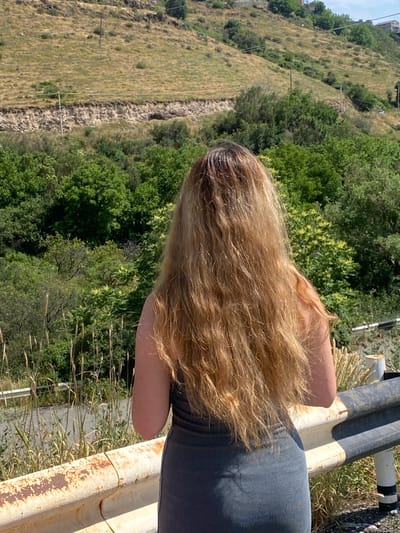 A woman with long, blonde, wavy hair is seen from behind, leaning against a rusty metal barrier. She is wearing a dark, ribbed top. The background shows a steep, dry, grassy hillside with scattered trees and some infrastructure, possibly a road or path. The scene appears to be outdoors on a sunny day, with abundant green foliage and dry grass suggesting a warm climate. The woman's posture and the setting evoke a sense of relaxation or observation. The location context provided is Ptghni, Armenia. There is no visible text in the image.