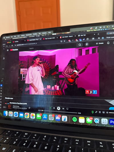A laptop screen displays a YouTube video featuring a musical performance, with elements of the surrounding room and keyboard also visible. The image is taken at an angle, presenting the laptop screen and its content rotated.
On the laptop screen, a video shows two individuals in what appears to be an indoor performance setting, possibly resembling an NPR Tiny Desk concert. One person, wearing a plaid shirt, plays an acoustic guitar. Another person, dressed in a white shirt and beanie, sings into a microphone, with several lit candles on a stand in front of them and a patterned backdrop behind. Pink or purple LED lighting frames the stage area. The "npr" logo is clearly visible in the top right corner of the video player. The video's title "Justin Bieber: Tiny D" is partially visible in a browser tab, and the URL `youtube.com/watch?v=Su6kidaGW_8` is displayed. The video is at the 0:15 mark of a 5:28 total duration. Below the video, text prompts to "Donate now" and "Give Back to Tiny Desk & NPR" are visible, along with a suggested video for "Ozzy's Song" by "Black Label Society, Zakk Wylde."
The laptop's Google Chrome browser has multiple tabs open, including "Inter black - Search," "New Tab," "YouTube," "Justin Bieber: Tiny D," "How To Add A FACE," "Ask G," and "Finish upda." The macOS dock is visible at the bottom of the screen, featuring numerous application icons such as Discord (with 2571 notifications), Mail, Safari, Messages (with 469 notifications), App Store (with 2 notifications), Settings, Chrome, Spotify, Notes, and QuickTime Player. The keyboard, partially in view, shows standard QWERTY keys and function keys F1 through F10. A light brown door frame from the room is visible in the background to the left of the laptop. The screen itself appears to have some smudges or dust.