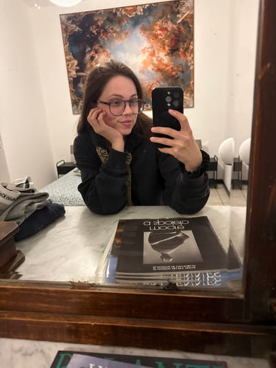 The image is a selfie taken in a mirror. A young woman with glasses is holding a smartphone, her face close to the camera. She is wearing a black sweatshirt with a Calvin Klein logo on the sleeves. To her right is a stack of books. One book has the title "a book about bloom" and another has "Bazzi" on the cover. Behind her, in the mirror's reflection, is a painting of an Italian Renaissance-style scene with figures and clouds. The bathroom counter is marble. An undergarment can be seen hanging from a drawer. The room contains a unique overhead lighting fixture. The photograph was most likely taken in the historical center of Perugia, Italy, since the artistic style of the painting in the mirror appears inspired by the artistic work displayed in the Galleria Nazionale dell'Umbria in Perugia.