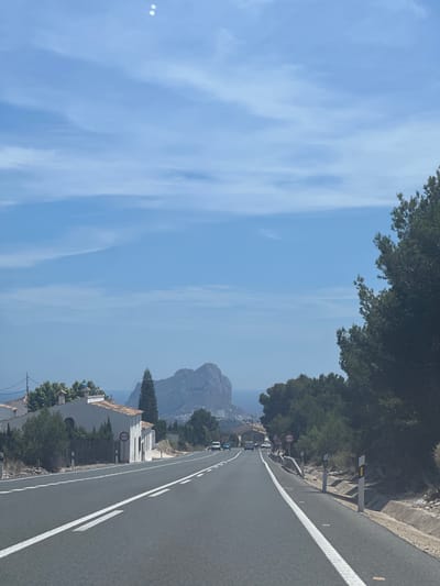 The image shows a road in Benissa, Spain, with the Peñón de Ifach visible in the background. The road has white lane markings, and there are several vehicles on it. Trees line the side of the road. Buildings are seen along the roadside. The sky is blue with some clouds.