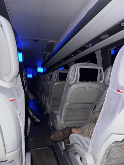 The image, rotated 90 degrees clockwise, captures the interior of a modern public transport vehicle, likely a long-distance bus or train. The setting is characterized by low light conditions, illuminated by distinctive blue ambient lighting along the floor of the aisle, suggesting night travel.
On the right side of the rotated image (representing the ceiling if corrected), light-colored panels or overhead compartments are visible. Below these, rows of passenger seats extend down the aisle. The seats are light grey or white and feature individual screens or tablets embedded in their seatbacks, indicating amenities for travelers.
In the upper-right portion of the image, the leg and foot of a person wearing a dark brown, lace-up boot are visible, resting in the seating area. Further along the dark floor of the aisle, another foot, clad in a black boot or shoe, can be seen.
The prominent blue LED lights, which appear as glowing rectangular or cuboid shapes, line the aisle, creating a contemporary and serene atmosphere. No discernible text is visible within the image itself. The scene depicts a quiet interior journey, presumably taking place in or near La Losa, Spain, according to the provided context.