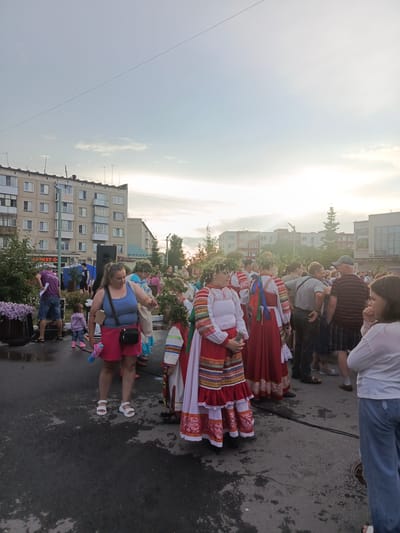 The image depicts an outdoor gathering of people in a city street, likely in Kryvodanovka, Russia, based on the metadata. In the foreground, several individuals are prominently featured. Two young women are dressed in traditional folk costumes, characterized by red and white embroidered dresses and leafy wreaths on their heads. One of them holds a microphone. To their left, a woman in a blue tank top and pink shorts is seen with a black purse slung across her body. She is holding a pink toy gun. Behind her, a young child in a pink hoodie and jeans is visible.
The background reveals a multi-story residential building with numerous windows, typical of Soviet-era architecture. Several other people are gathered in the street, some in casual attire, and others appearing to be part of the same cultural event. The street surface is asphalt, with visible wet patches, suggesting recent rain or cleaning. Trees and bushes are present along the side of the street, and a large speaker system is set up. The sky is overcast with a bright horizon, indicating either early morning or late afternoon. Various signage is visible on the buildings, including what appears to be a store sign with Cyrillic lettering.