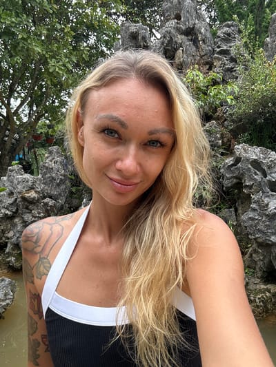 A blonde woman takes a selfie outdoors. She is wearing a black and white swimsuit. Behind her are large rock formations and trees. The woman has tattoos on her left arm and shoulder. The location is identified as Thôn 2, Vietnam.