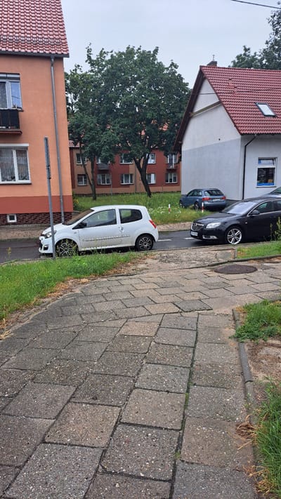 This is a medium-shot, eye-level, outdoor photograph taken on a cloudy day. The scene depicts a street in the city of Krosno Odrzańskie, Poland. In the foreground, a paved walkway made of large, grey, interlocking concrete slabs extends towards the right of the frame, bordered by green grass and some dirt on the right. Behind the walkway, a street with a white line marking is visible, with two cars parked on it. The car closest to the foreground is a dark grey, four-door sedan. To its left is a white, two-door hatchback car. Further down the street, a blue four-door sedan is parked.
The background features a red-tiled roof of a residential building on the right, with a white and grey facade. To the left, an orange-colored building with multiple windows and a small balcony is visible. Several trees with dense green foliage are situated between the buildings, providing shade. The overall impression is a residential street in a European town.