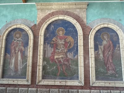 The image shows a religious mural on a wall, depicting three arched panels. The mural is located in the Church of Saint Demetrius in Sremska Mitrovica, Serbia.
The left panel depicts a saint in red and white robes, holding a book and wearing a crown. The central panel features a warrior saint in armor, holding a spear and a shield, with a red cloak. The right panel shows a female saint in a red robe, holding a cross. All three figures have halos, indicating their sainthood. The background of each panel features a stylized landscape with buildings.
The mural is framed by arched brickwork and stone. The wall above the mural is painted a turquoise color. The bottom of the image shows a brick base with carved patterns. The lighting suggests an indoor setting, likely a church interior.