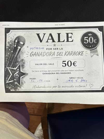 This is a photograph of a certificate or voucher awarding someone named KATHERINE for being the winner of a karaoke event. The certificate has a graphic of a microphone and a star. The text on the certificate reads: "VALE", "KATHERINE POR SER LA GANADORA DEL KARAOKE", "VALOR DEL VALE 50€", "Se hace entrega del presente vale por haber resultado GANADORA DEL KARAOKE.", "FECHA: 12 / 06 / 2026", "FIRMA Y SELLO:", and "¡Enhorabuena por tu merecida victoria!". The certificate is signed with blue ink. The background of the image appears to be a dark, possibly textured surface. The timestamp in the upper left corner indicates "12/6/26, 23:36", and a "Gmail - (no subject)" label is visible at the top. The bottom portion of the image shows the edge of a white paper with some text, and below that, a dark surface which could be a table or floor, with parts of clothing or fabric visible. There is no identifiable information within the image that places the scene in Torrejón de Ardoz, Spain.