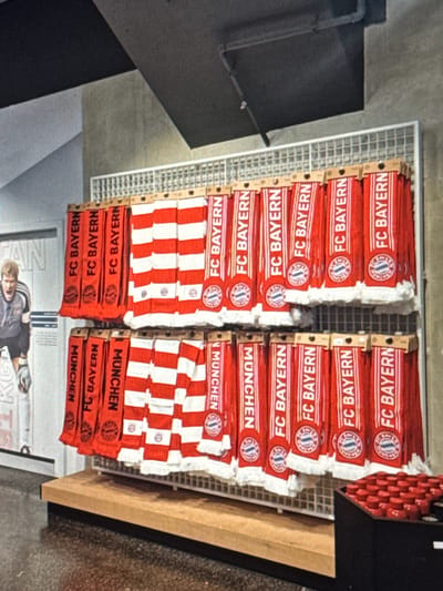 The scene depicts an indoor retail environment located in Dehqonbozor, Uzbekistan. A two-tiered white metal grid display rack occupies the center-right of the frame, holding numerous football club scarves. The scarves are predominantly red with white lettering, featuring "FC BAYERN" and "MÜNCHEN" text, along with the FC Bayern Munich logo. Some scarves exhibit a red and white striped pattern. They are neatly folded and suspended from cardboard tags. The display rack rests on a light brown wooden base.

To the left, a large, light-colored poster or mural is visible on a grey wall, featuring a fair-haired male figure in a grey long-sleeved shirt and goalkeeper gloves, posed with an open mouth. Partial text, including "AN," is visible on this poster. The background reveals a dark grey or black ceiling with exposed pipes or conduit running across it. The floor is dark and reflective. In the lower right corner, a black container holds multiple small, uniform red cylindrical objects, arranged upright.