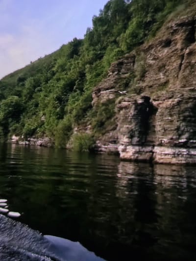 The image depicts a landscape with a rocky cliff face covered in lush green vegetation on one side, and a dark, reflective body of water on the other. The water mirrors the cliff and foliage, creating a distorted, rippled effect. The sky is a pale blue, suggesting daytime. The overall impression is one of a natural, tranquil scene, possibly a river or lake bordered by a steep, overgrown embankment. There are no visible people or man-made objects, and no text is present. The image's orientation is unusual, with the sky appearing at the bottom and the water at the top, indicating it may have been taken from a low angle or that the image itself has been rotated. Given the location context of Dubno, Ukraine, this could be a scene from a Ukrainian natural reserve or a picturesque riverside. The lighting suggests it is daytime, likely late morning or afternoon, with the reflective water capturing the ambient light. The weather appears to be clear and calm.