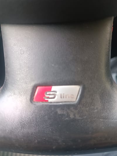 This is a close-up, vertical shot of the steering wheel of a car, specifically focusing on the "S line" badge. The steering wheel appears to be made of black, textured material, possibly leather. The "S line" badge is rectangular with rounded corners. It features a red and silver design. The left side of the badge is a solid red area, while the right side is silver. The silver area has a stylized "S" embossed on it, with the word "line" written in a simple, sans-serif font below and to the right of the "S". The badge is slightly inset into the steering wheel. The lighting suggests it is daytime, and the image appears to have been taken inside the car, possibly in a parking lot or on a street. There are no people visible in the image, and the focus is solely on the steering wheel detail. The overall impression is of a close-up detail shot highlighting a specific trim level of the vehicle.