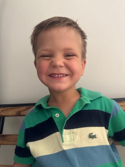 A young boy is smiling with his eyes squinted shut, revealing gaps in his front teeth. He is wearing a striped polo shirt with green, navy blue, and light blue bands, and a small green crocodile logo on the left side of his chest. The boy is positioned in the foreground of the image, with a plain, light-colored wall behind him. The overall scene appears to be indoors. The image does not provide any information about the city of Les Rousses, France, or its specific location within the city.