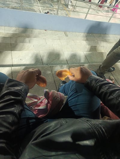 The image shows a first-person perspective of someone sitting on concrete steps, possibly stadium seating, holding a pastry-like food item in each hand. The person is wearing jeans and a black leather jacket, with a pink and white bag visible between their legs. Another person's legs are visible to the right. Above the person, there is a metal railing with a concrete wall. There are other people standing/walking above the wall. The location is identified as El Cisne, Ecuador.