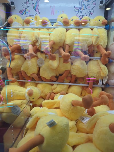 A medium shot captures a claw machine filled with numerous yellow plush duck toys, each featuring orange feet and beaks, and some wearing small white bibs. The toys are densely packed across multiple levels, with some hanging by their legs and others slumped over. The machine's interior is lit, revealing the reflective glass exterior, which shows distorted reflections of the toys and surrounding environment. The background features a colorful graphic of a unicorn and rainbow, with the word "Worry" partially visible in pink lettering. The scene is located within Xishan District, China.