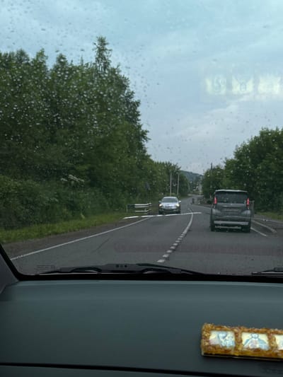 The image is taken from inside a car, looking forward through a rain-streaked windshield. The road ahead is wet, with white dashed lines marking the lanes. Two cars are visible on the road: a silver sedan in the distance and a dark grey SUV closer, in the right lane. Both cars appear to be moving forward. The surrounding environment is lush with green trees and vegetation on both sides of the road. The sky is overcast and appears to be a light grey color, suggesting a cloudy or rainy day. In the foreground, on the dashboard of the car, is a religious icon consisting of three small framed images. The scene appears to be a rural or semi-rural road. The location is described as the city of Kurtukovo, Russia.